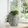 Buddha’s Head planter with lush indoor plants sits on a light wooden table by a window, creating a peaceful display with greenery.