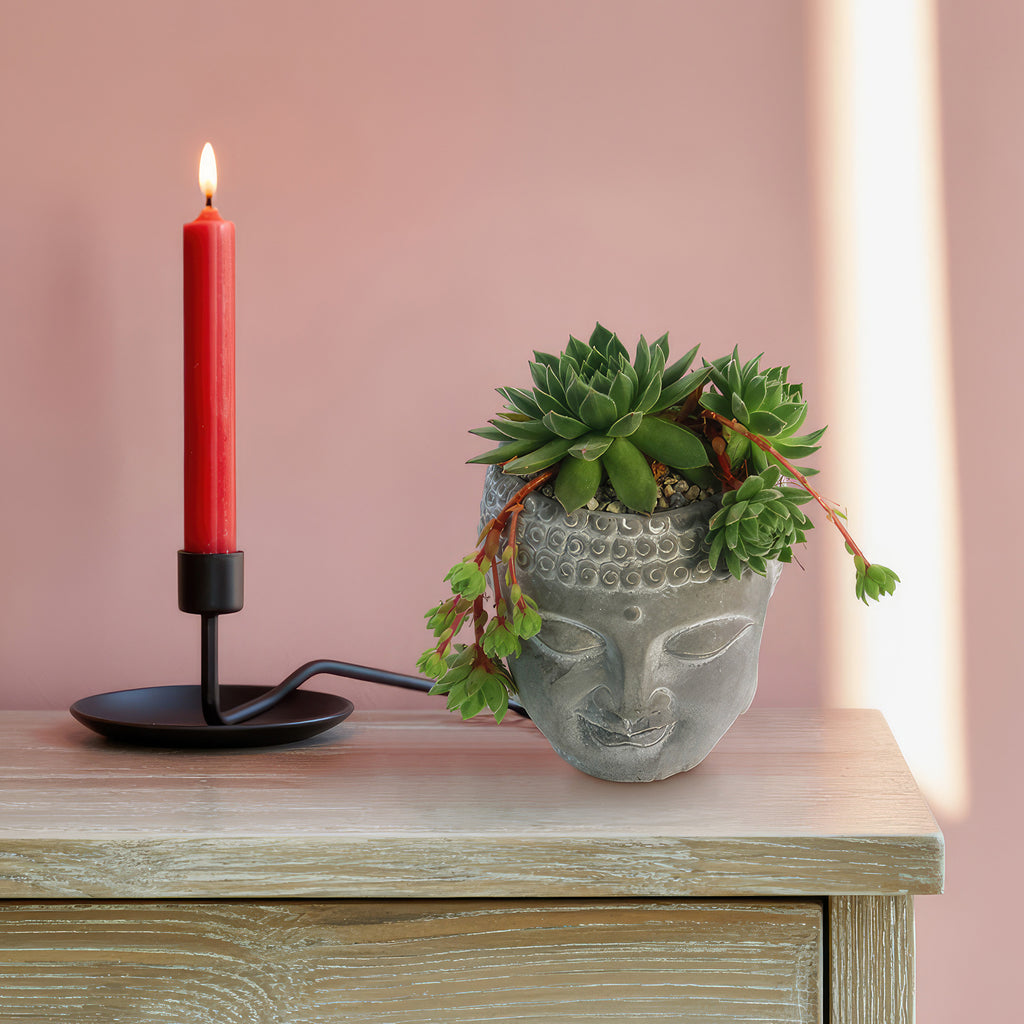 Buddha’s Head planter with live indoor plants on a light wood table beside a red candle in a black holder, set against a pink backdrop.