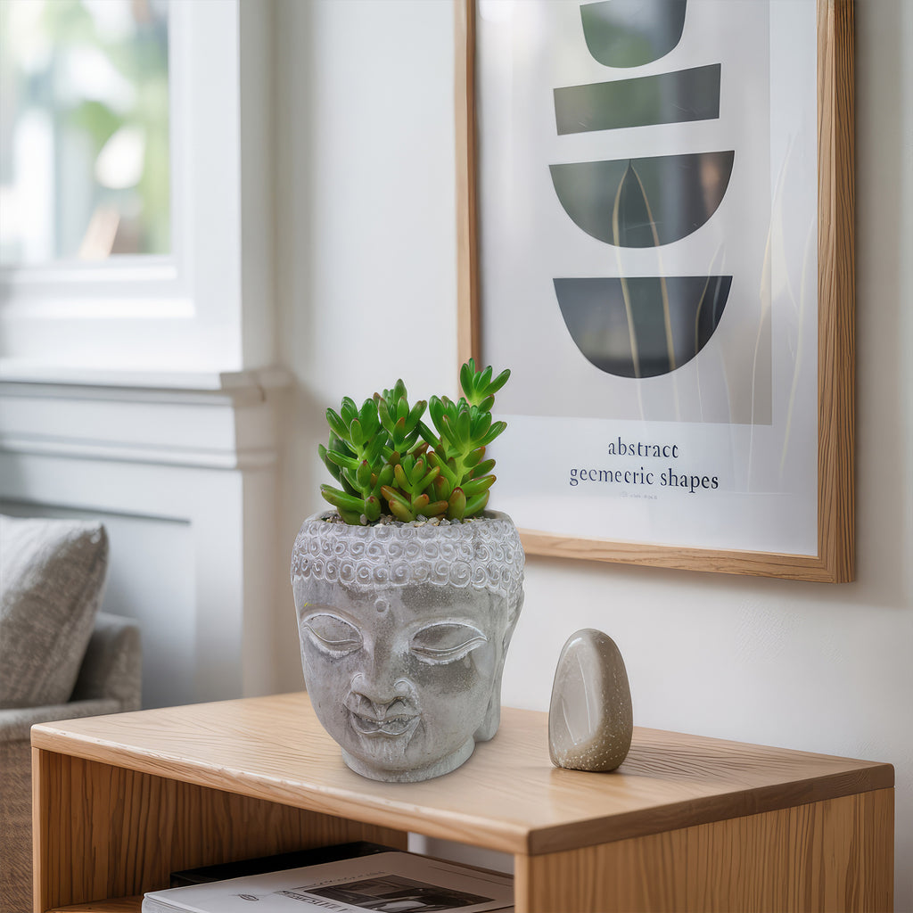 A Buddha head planter with live indoor plants on a wooden table beside a smooth stone, set against abstract art for a calm, stylish look.