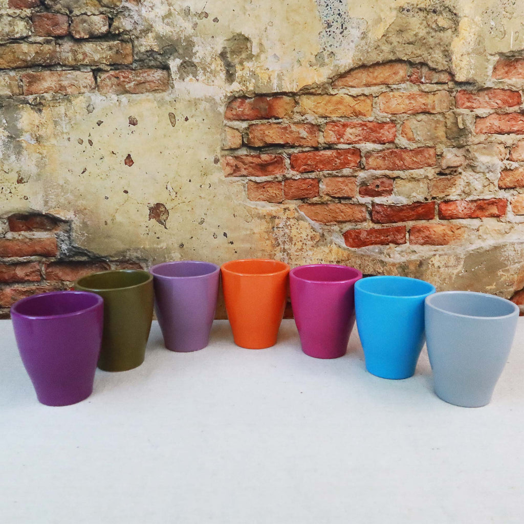 Seven glossy ceramic pots in seven colors sit on a white surface before a brick wall, perfect as decorative or indoor plant pots.