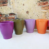 Four glossy ceramic pottery pots in purple, olive green, light purple, and orange on a light surface with a rustic brick wall backdrop.
