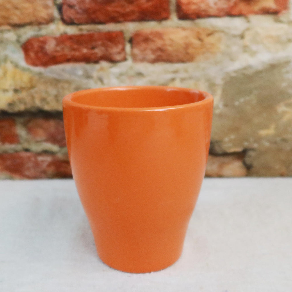 An orange ceramic glossy pottery cup sits on a white surface, with a blurred brick wall and decorative planters in the background.