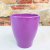A vibrant purple glossy ceramic planter sits on a light surface, with a blurred stone and brick wall visible in the background.