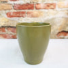 Glossy olive green ceramic pottery cup with a planter-inspired design on a white surface, brick wall blurred in the background.