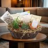 Gift basket with two small potted succulents, gardening tools, care cards, a white candle, and straw on a wooden table in a cozy living room.